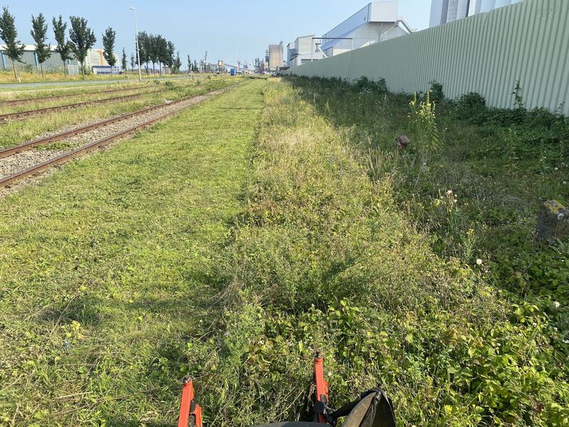 Ruwterrein maaien langs spoorrails op een industrieterrein, met een gemaaide strook naast hoog opgeschoten gras